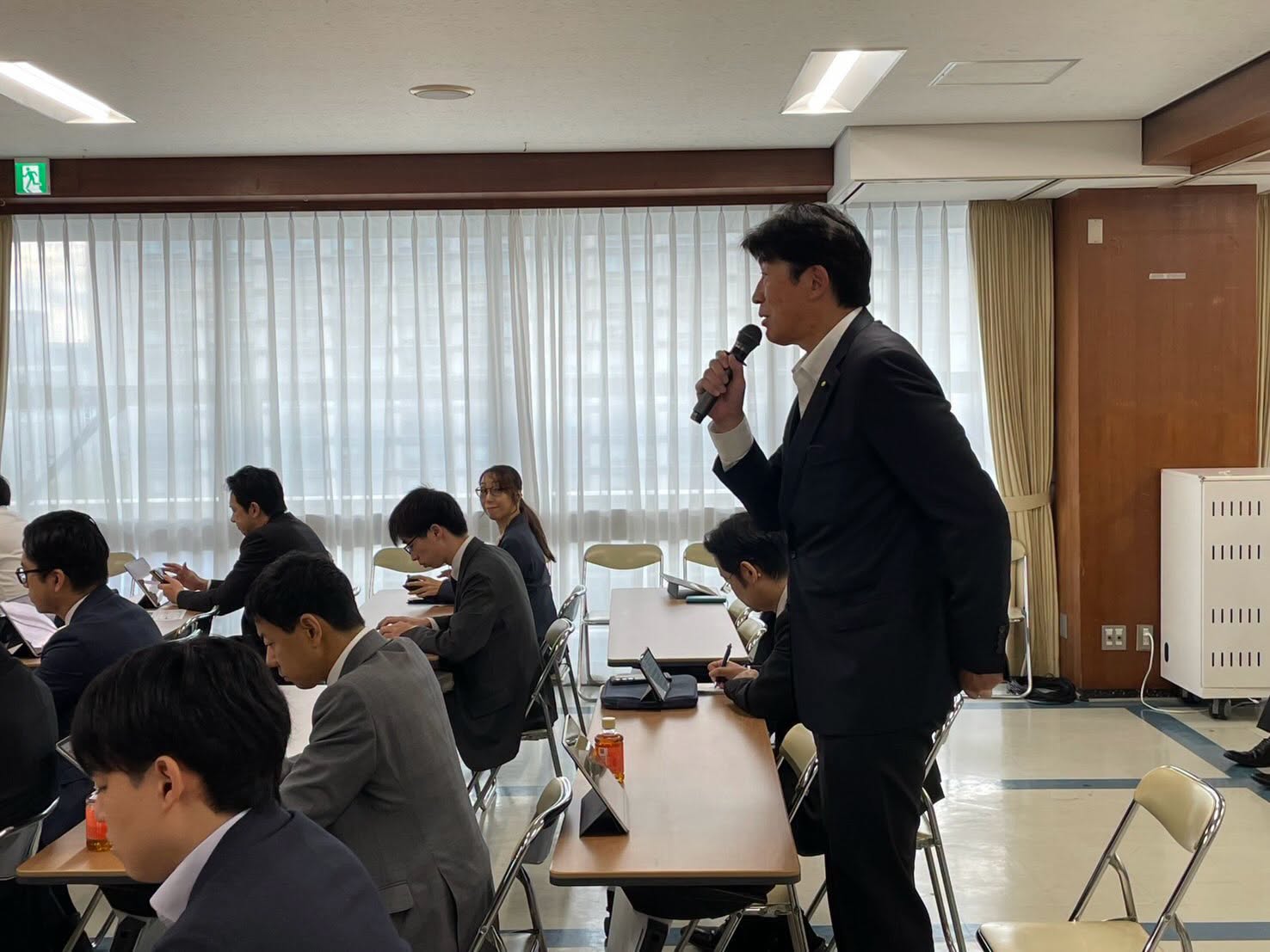 会議でマイクを持ち発言する様子。窓側の席から撮影