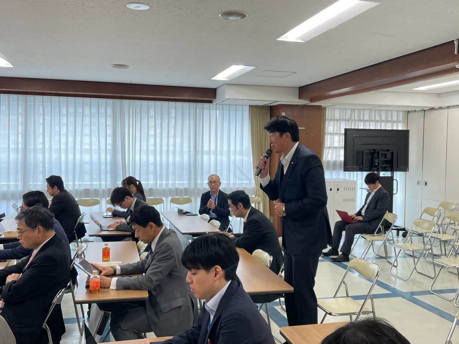 会議でマイクを持ち発言する様子。会場を広く写した別角度の写真